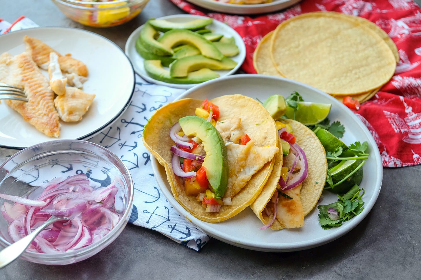 Plate of sliced avocado, plate of cooked whitefish, small bowl of sliced red onion, and two whitefish tacos displayed on a table.