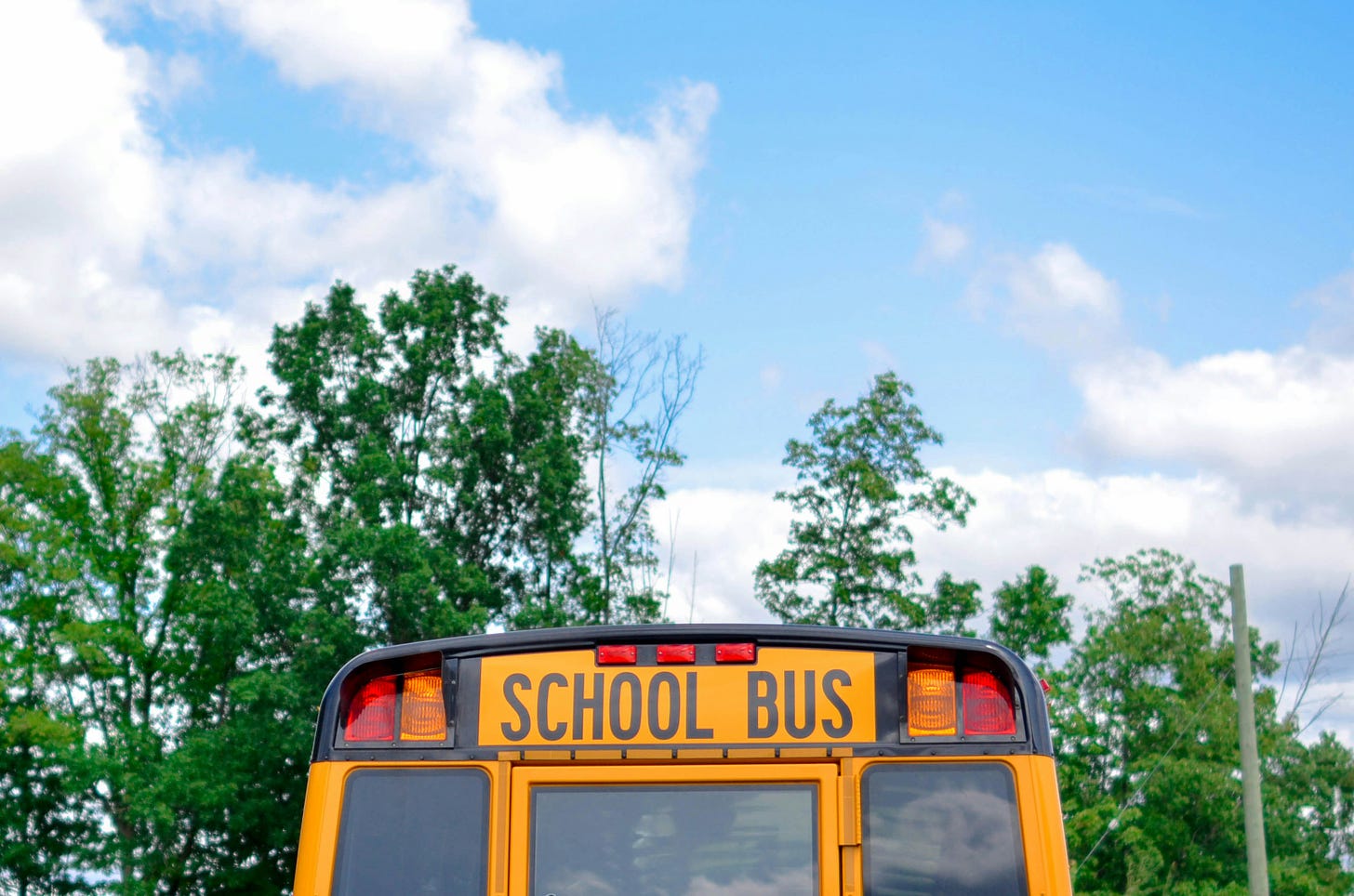 The back of a yellow school bus set against green trees and a partly cloudy blue sky.