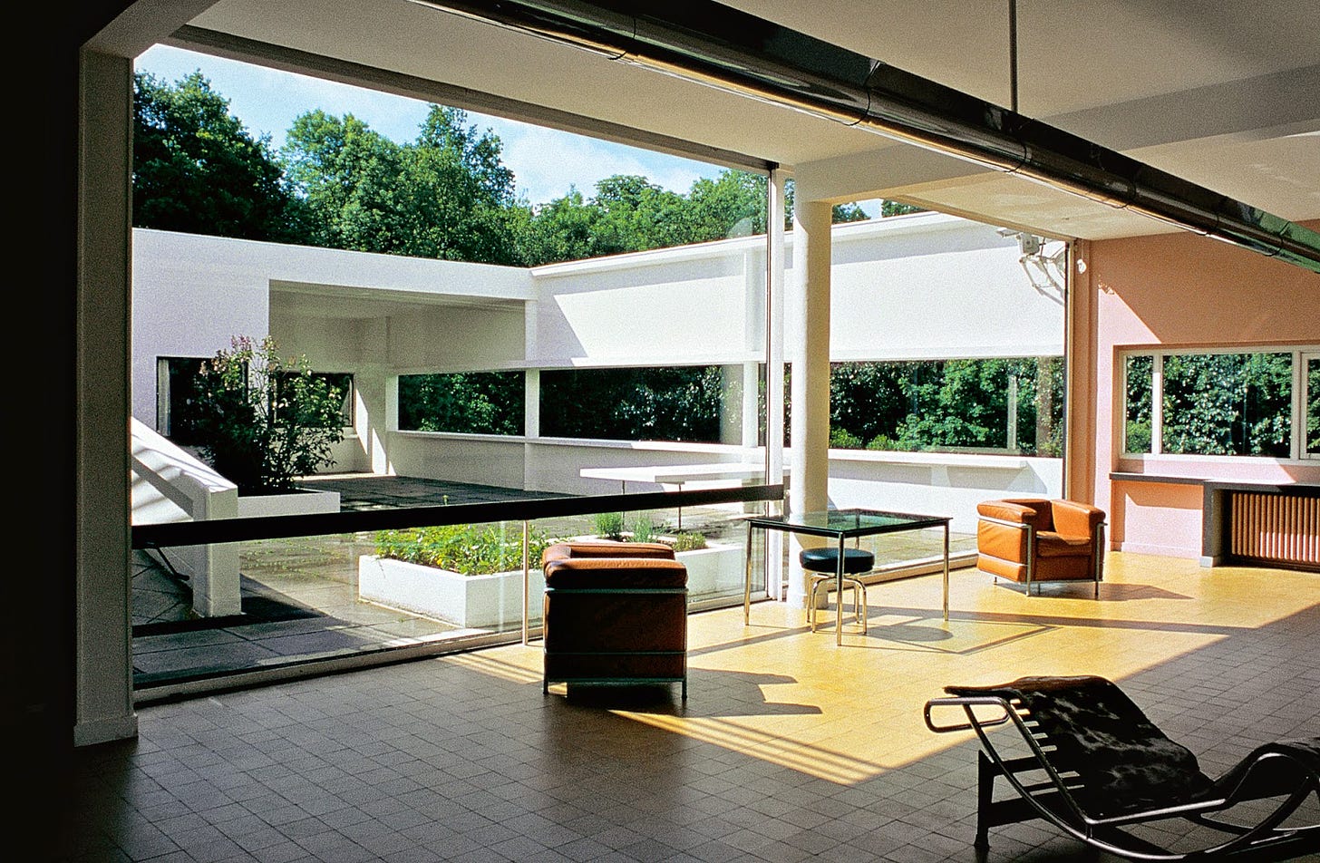 Sunlit seating area in a building with wide open walls/windows, surrounded by trees