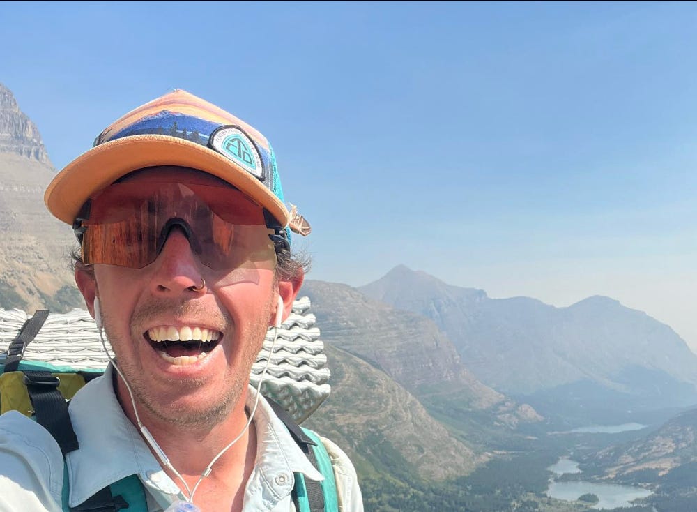 photo of Marlin Sill from TTFA Episode "The Trail." Marlin is taking a selfie while standing on the top of a mountain. He has a big smile on his face and is wearing sunglasses, earbuds and a baseball cap. On his back is a very heavy-looking backpack.
