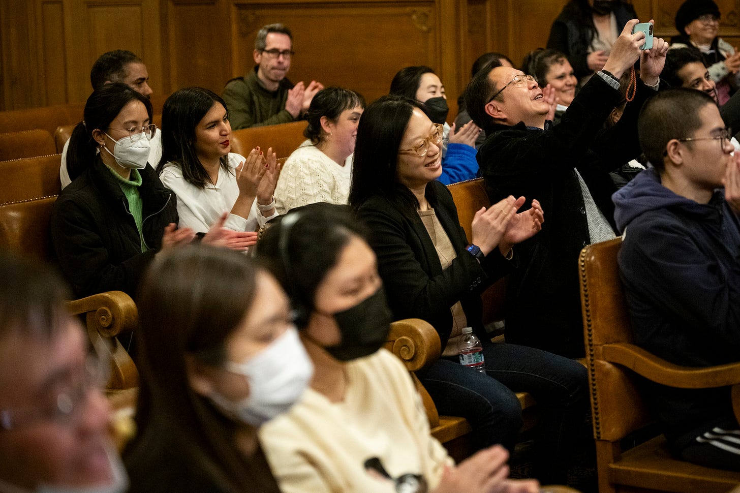 A group of people with some wearing masks, applaud in a room.