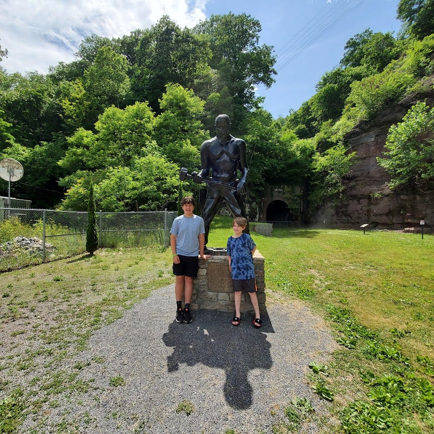 Two white boys stand in front of a statue of John Henry under a blue sky, surrounded by trees in full leaf. 