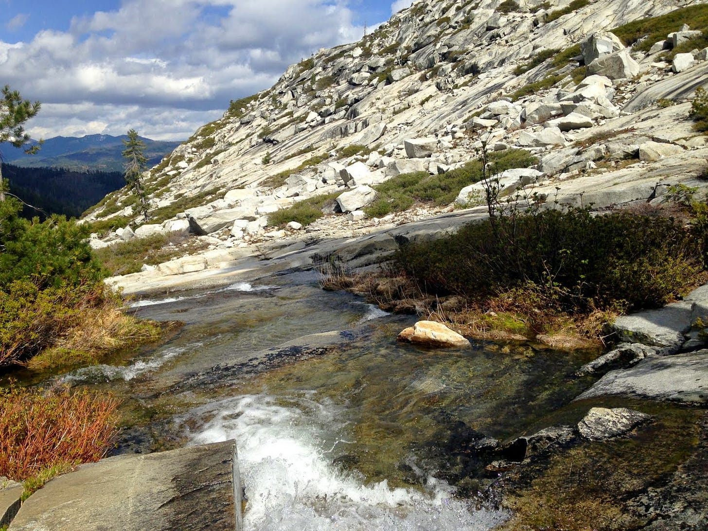A shallow stream runs down the side of a mountain composed of blocky white rocks with mountains and evergreen trees off into the distance.