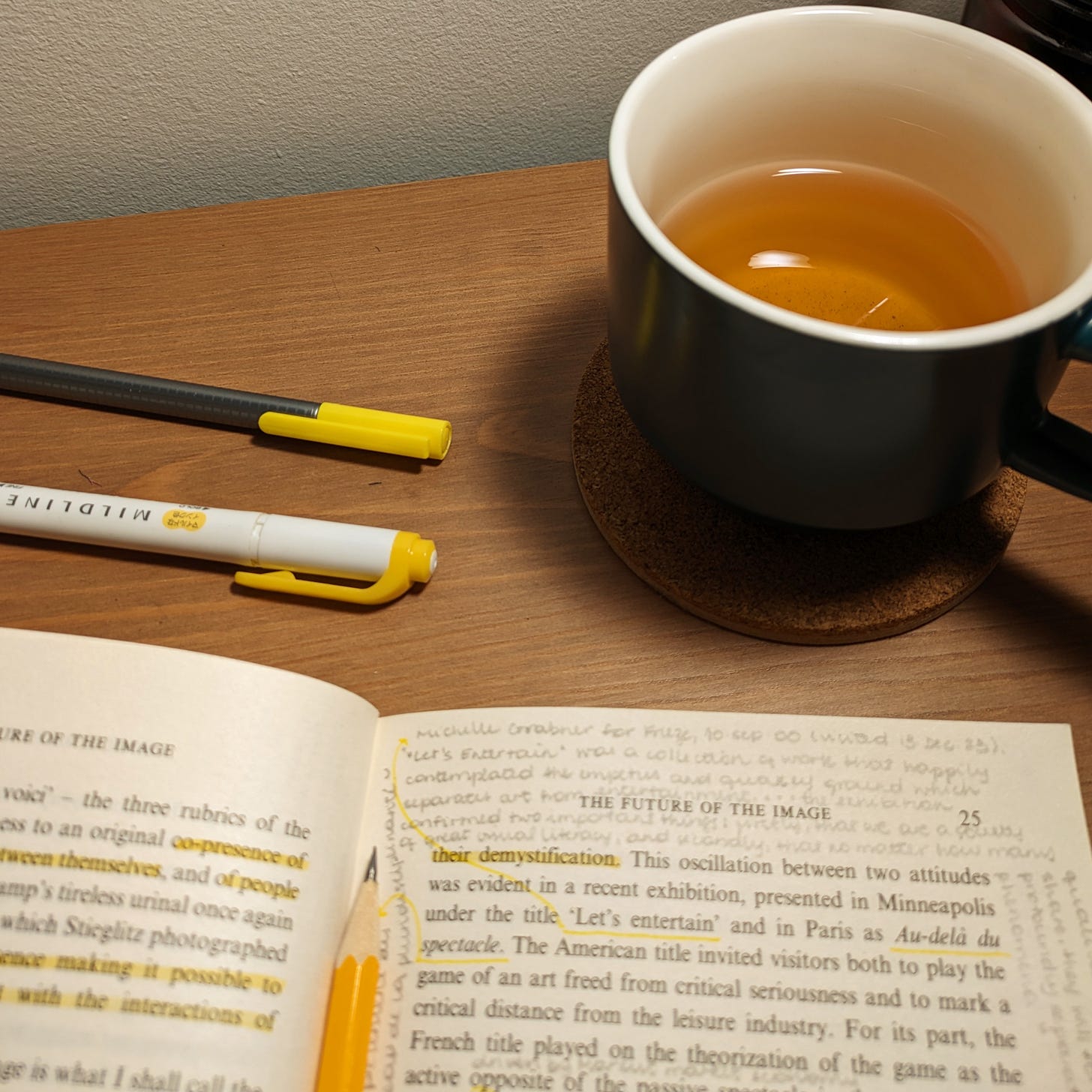 Photo of a desktop, with a dark green mug of tea at top right, a yellow fineliner pen and a yellow highlighter on the desk, and a book open with annotations across the top of the right-hand page with a yellow pencil holding it open in place.
