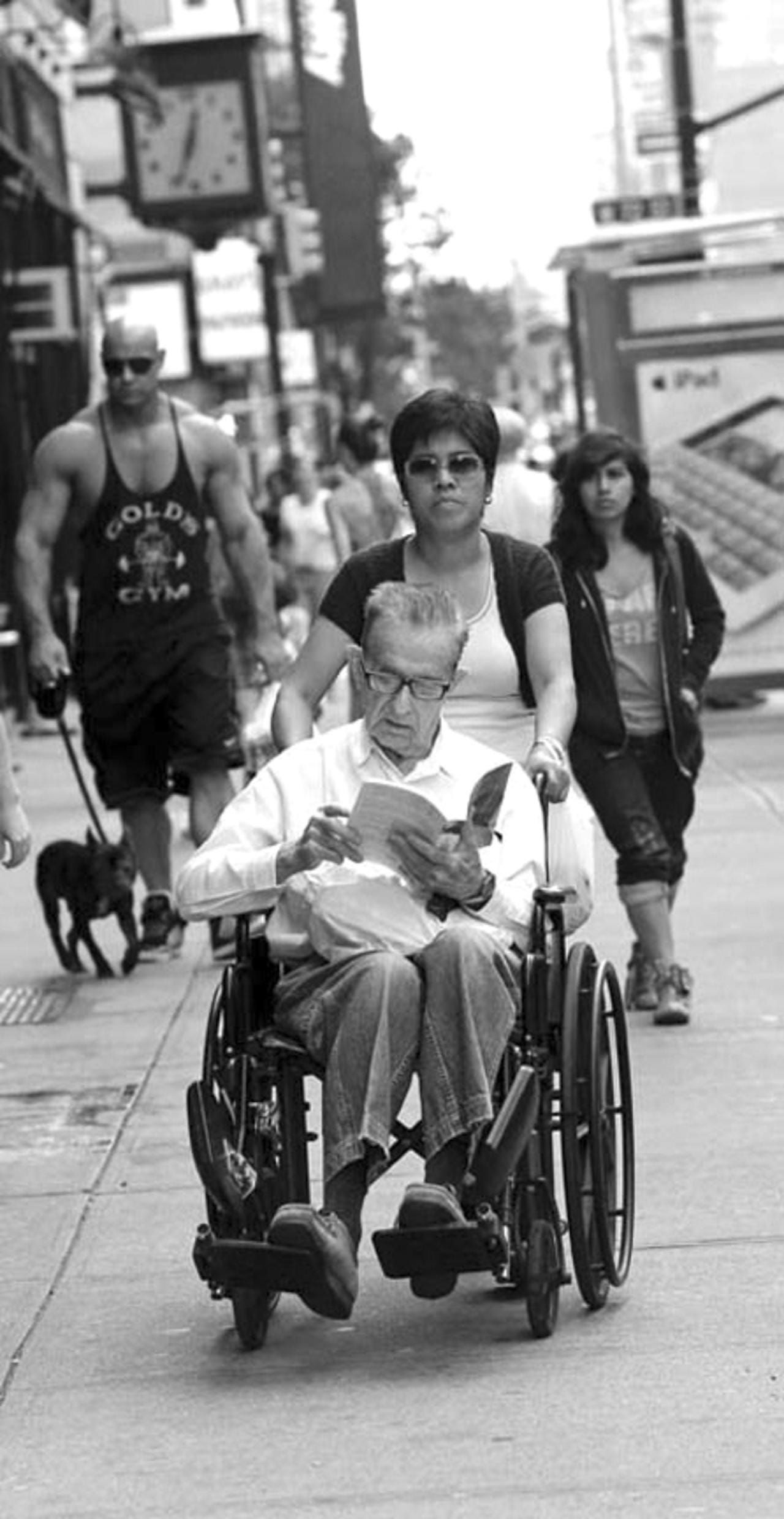 A man in a wheelchair reads a book while a bodybuilder walks his dog behind him on Amsterdam Avenue.