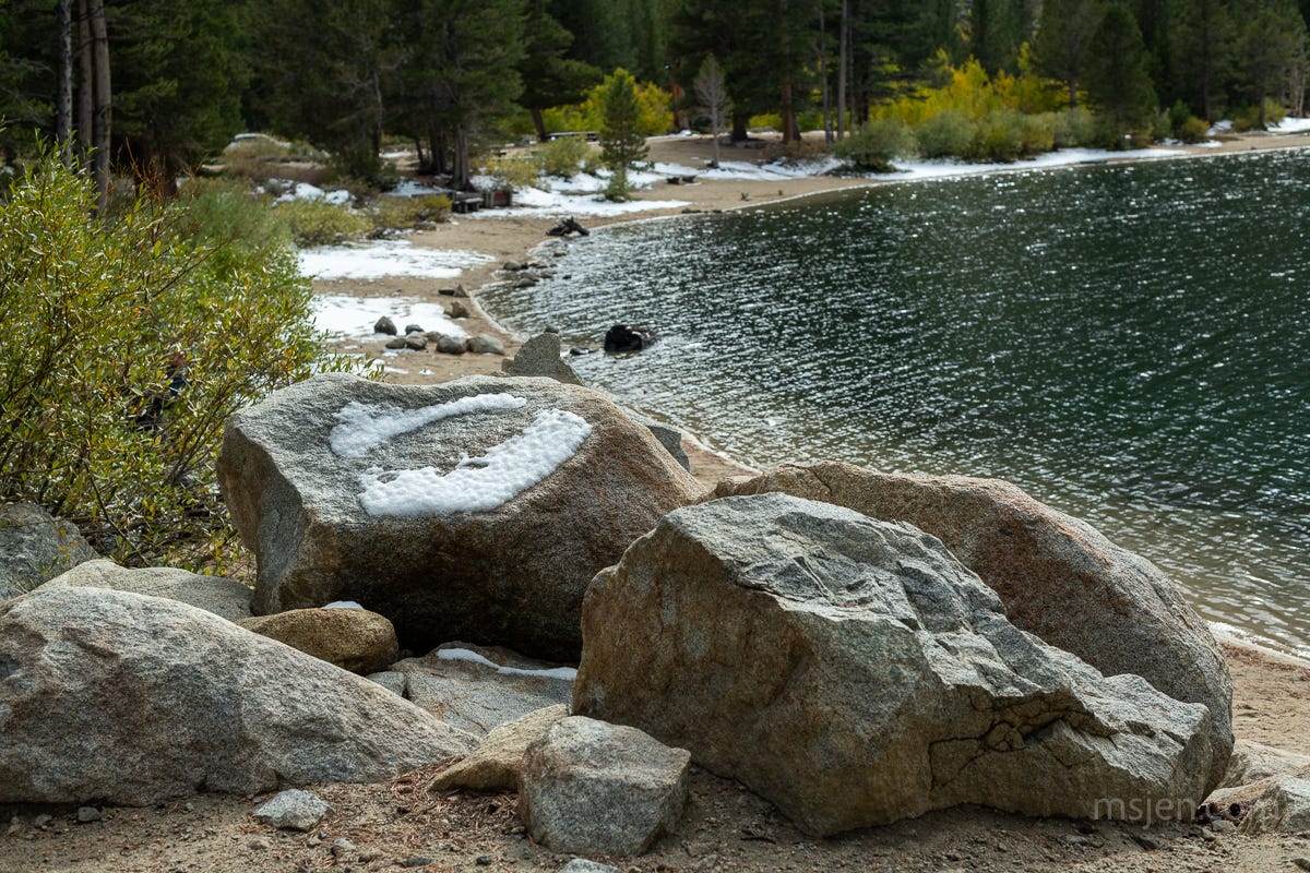 People sized large rocks with circles of patchy snow on them in the foreground, and the edge of Rock Creek Lake with more snow lining the shore in the background.