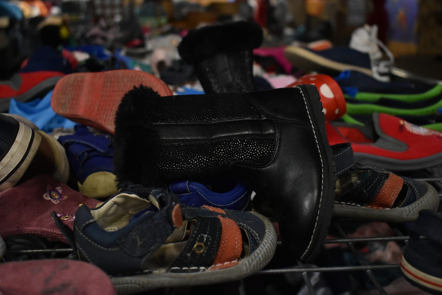 Foto de un mostrador en una tienda de artículos reciclados  y de segunda mano, que muestra un apilonamiento de zapatos de niños. En primera plana hay unas boticas negras y unas sandalias.