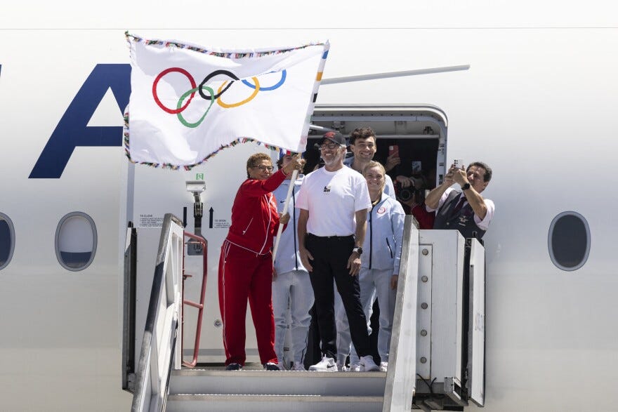 L.A. Mayor Karen Bass waves the Olympic flag as she exits a plane. She's surrounded by several people and wearing a red track suit.