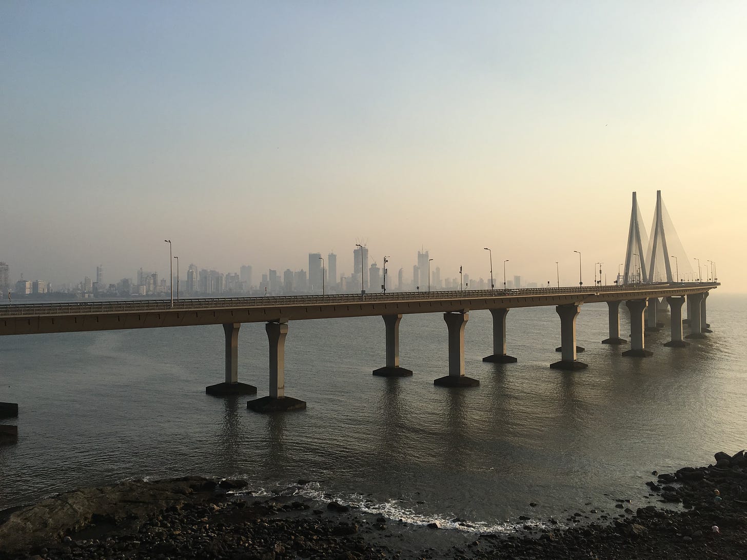 View of the Bandra-Worli Sea Link, with the pillars seen embedded into the ocean. The sky is hazy with a setting sun. In the background, the Mumbai skyline is faintly visible. 