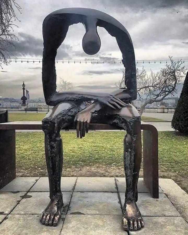 A bronze sculpture in a park. It is a large, somewhat abstract, sculpture of a person sitting on a bench. The sculpture has their head bowed, hanging down in a defeated position. Where the torso should be is instead a large, hollow square so that you can see right through the sculpture. 