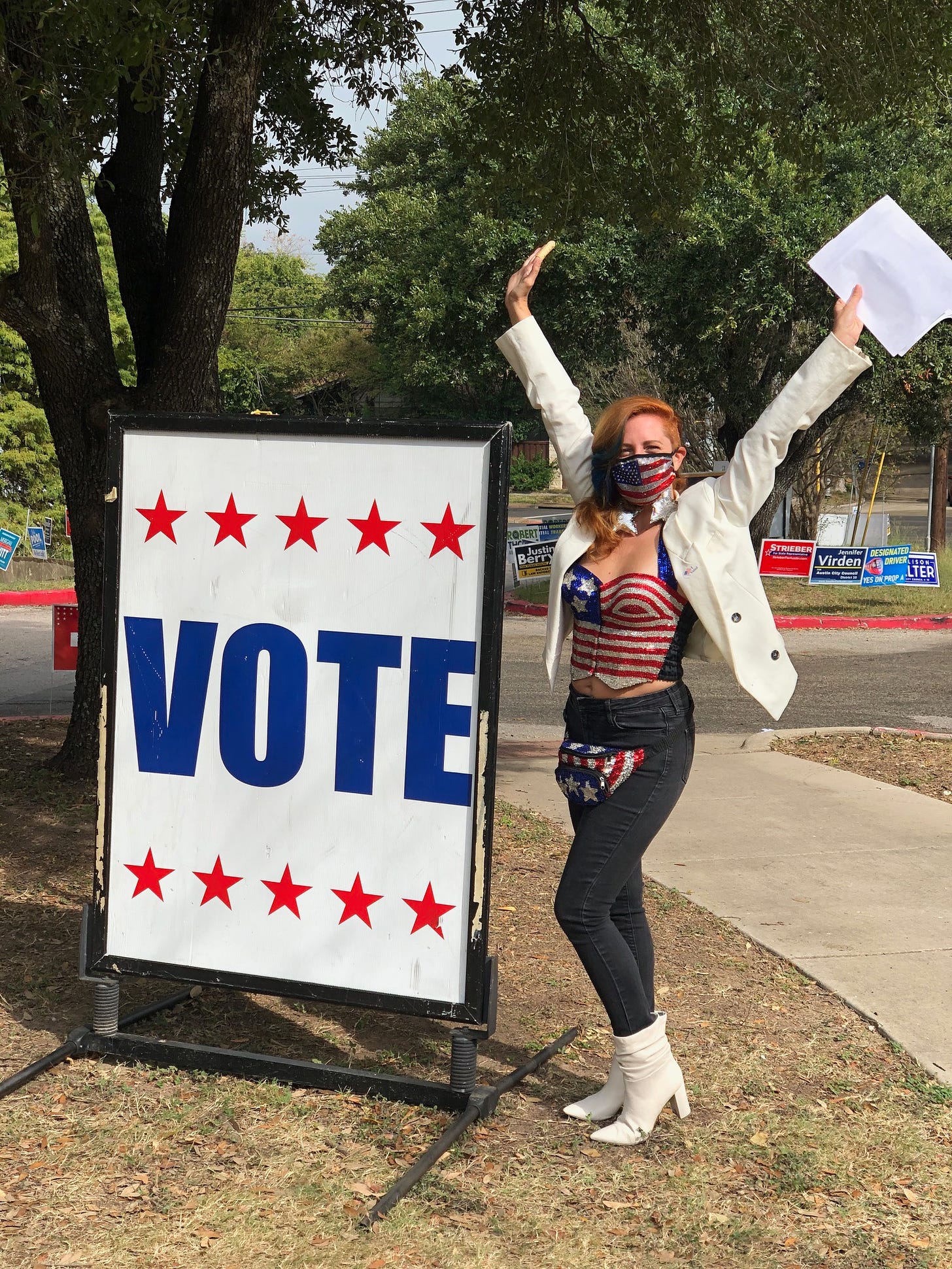 Becky wearing a white blazer over a sequined American flag bustier and raising her hands in a V. She is next to a giant VOTE sign outside a polling location.