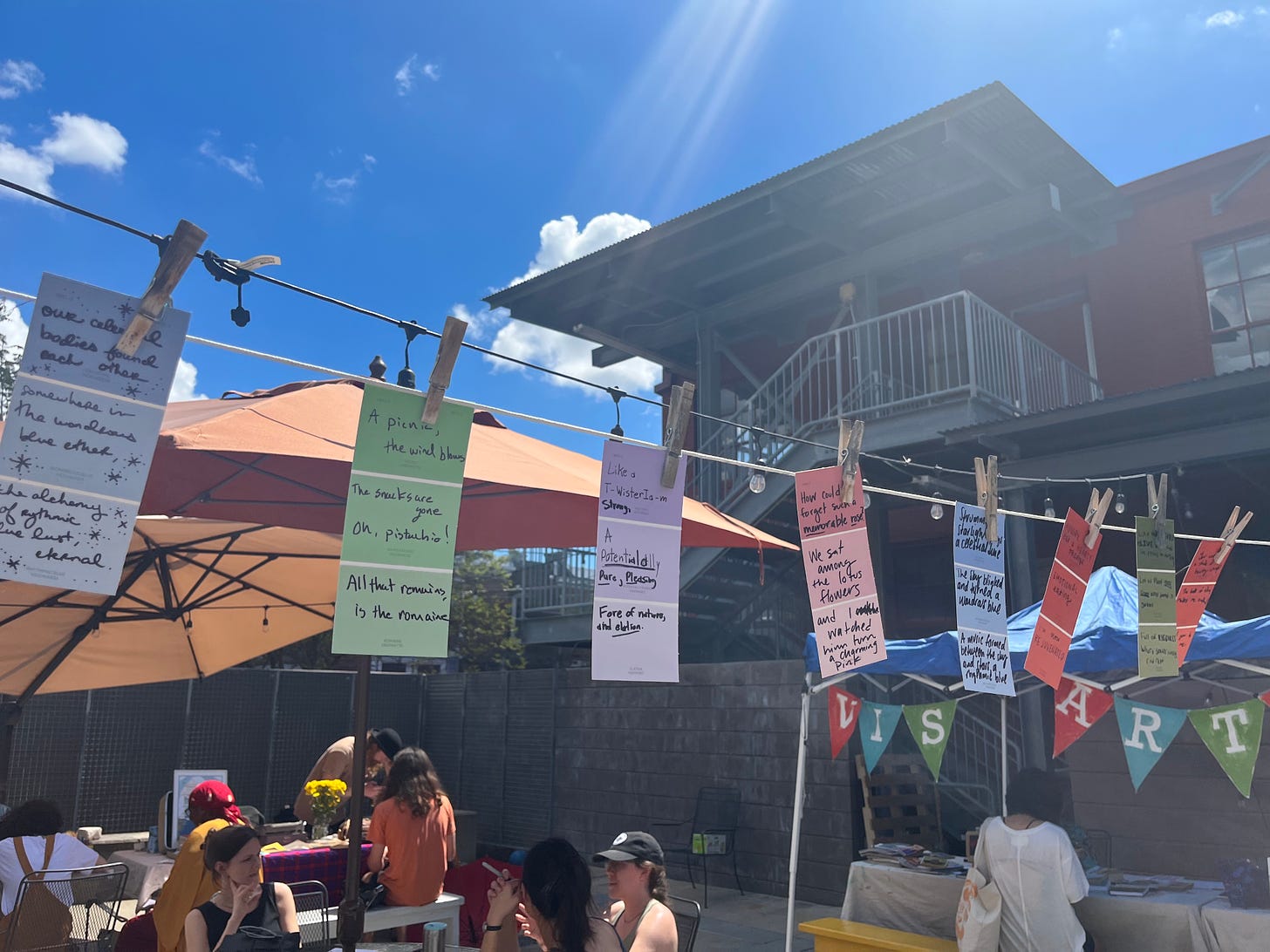 Photo of multi-colored paint chips with poems written on them hanging across a laundry line. A blue sky with the sun shining above, and people sitting at tables doing activities below.