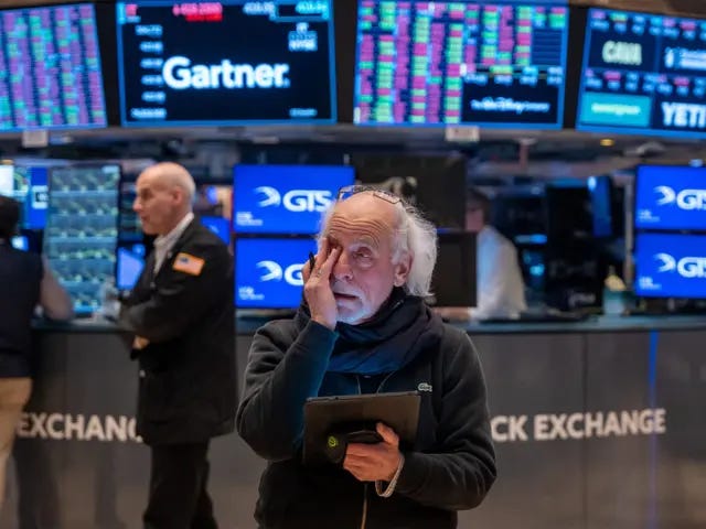 New York Stock Exchange trading floor with multiple monitors displaying stock market data. In the foreground a trader has his hand to his face.