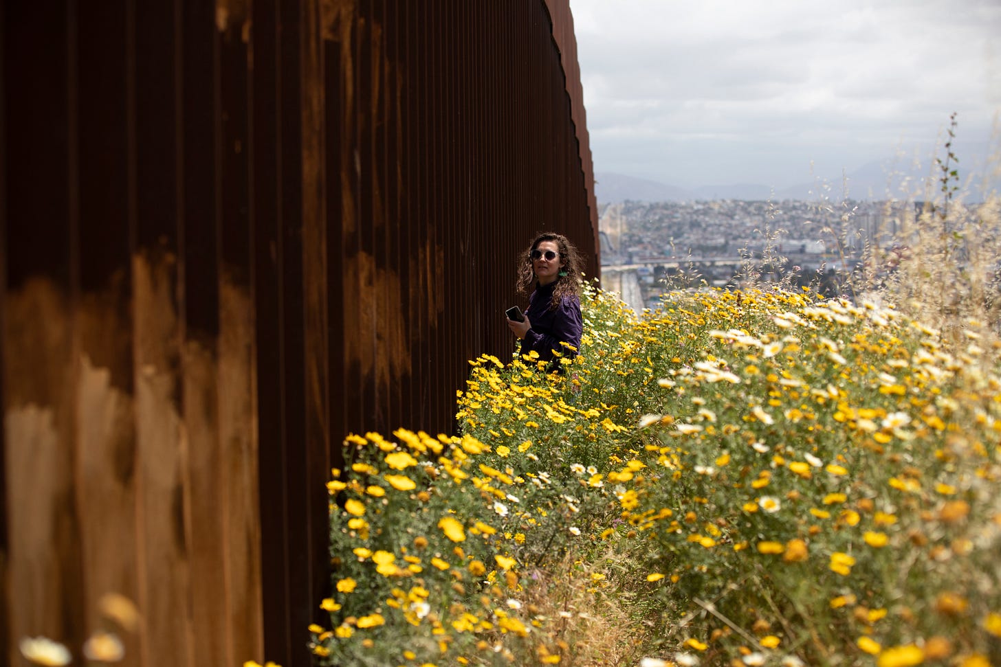 A woman journalist stands next to a tall wall with spring flowers blooming behind her