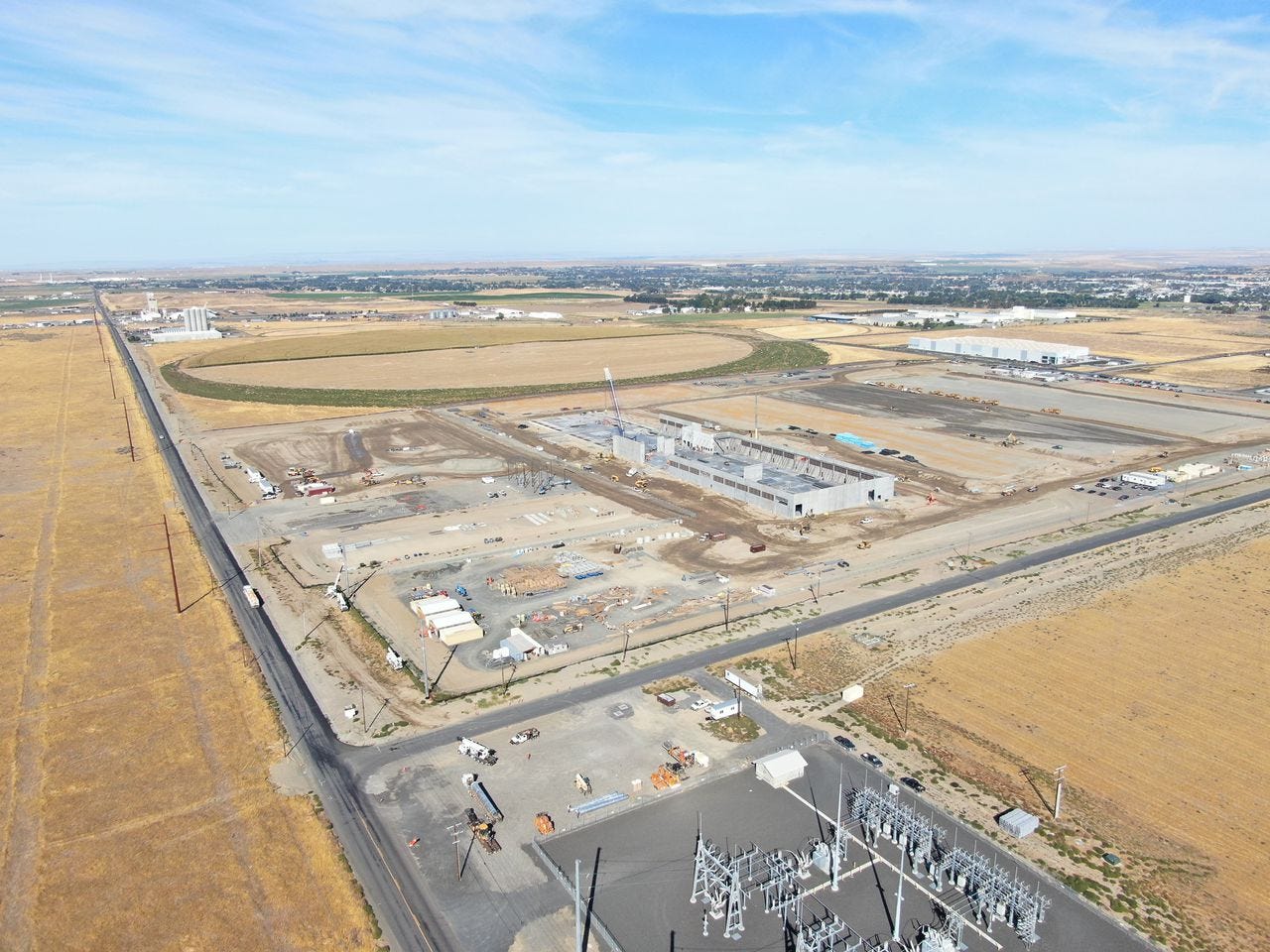 A concrete building with a large crane under construction. A circular agricultural field is beyond it; an electrical power substation is in the foreground. A concrete building with a large crane under construction. A circular agricultural field is beyond it; an electrical power substation is in the foreground.