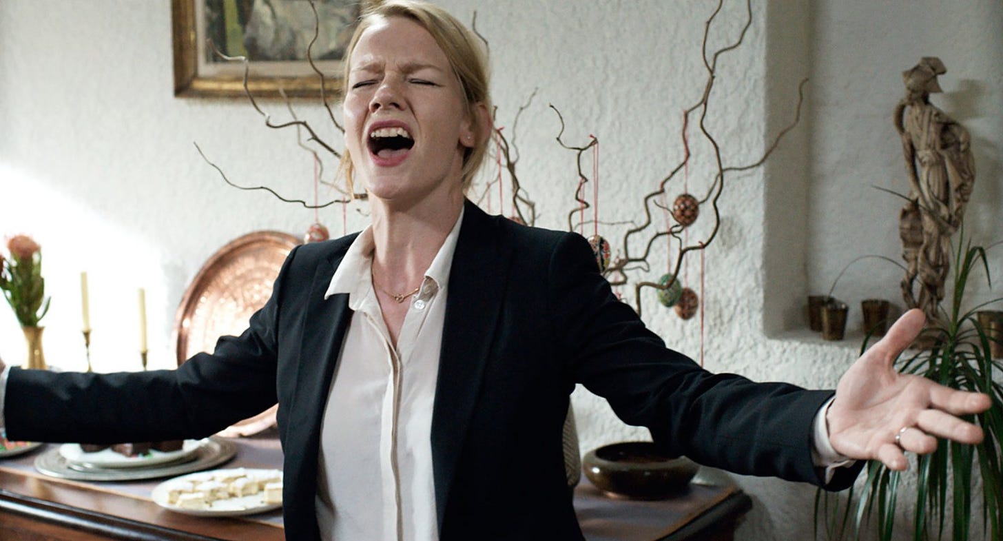 A film still of a woman in a black blazer and white top standing in a dining room area. She closes her eyes and has her mouth open wide, like she's singing. She raises her hands by her sides. 