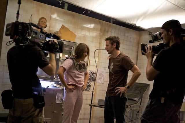 A behind-the-scenes photo of Scream Queens, with a contestant and a director discussing a shot while being filmed on camera. They're in a grimy-looking hospital room set.