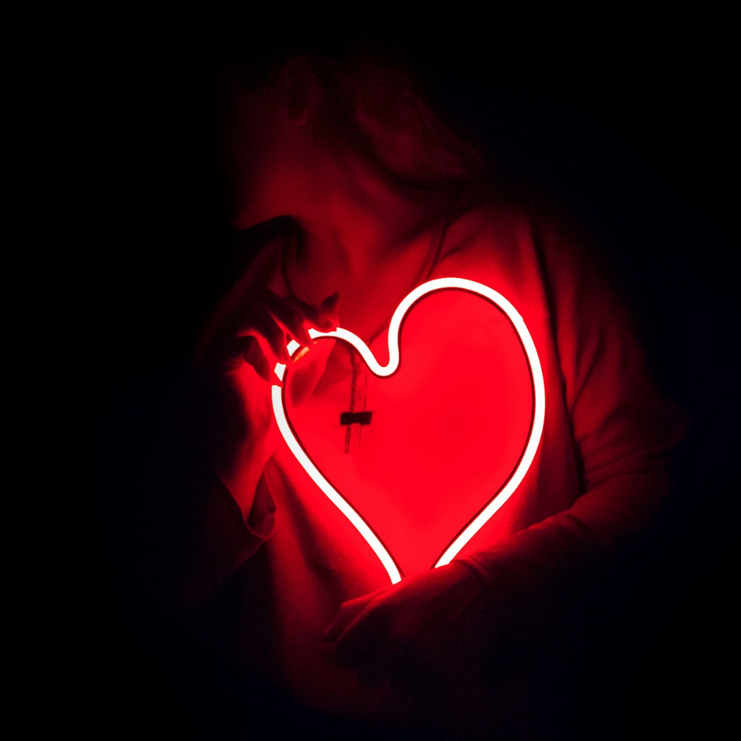 In the darkness, a person in a hoodie hugs a heart-shaped, soft red neon light close to their body. In the darkness, a person in a hoodie hugs a heart-shaped, soft red neon light close to their body.