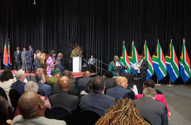 Line of people look at a man wearing a green judge's robe, standing in front of a row of flags, while seated people look on