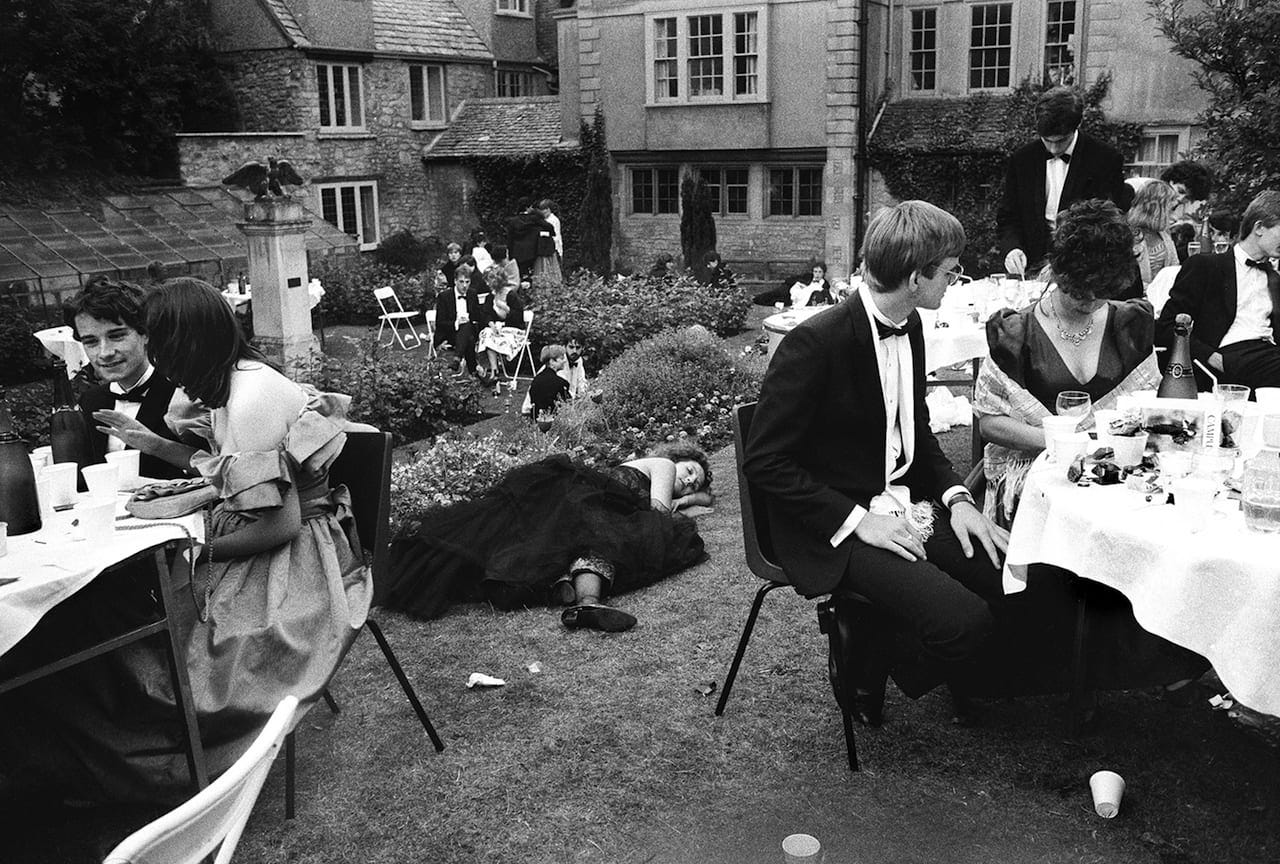 Black and white photograph taken on a lawn outside a large, mansion-like structure. In the center of the image, a young woman in a black gown sleeps peacefully on the grass. To the sides of her, groups of elegantly dressed young people sit around white clothed tables covered in wine glasses and white cups. 