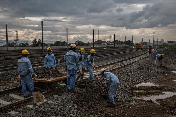 Workers in blue uniforms shovel and collect rocks along a train track.