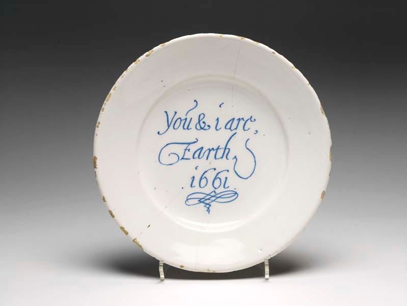 A tin-glazed earthenware plate with a hard white glaze, slightly bevelled rim, shallow-well and central inscription in blue: 'You & i are/ Earth/1661' with a paraph underneath (a flourish, usually at the end of a signature).