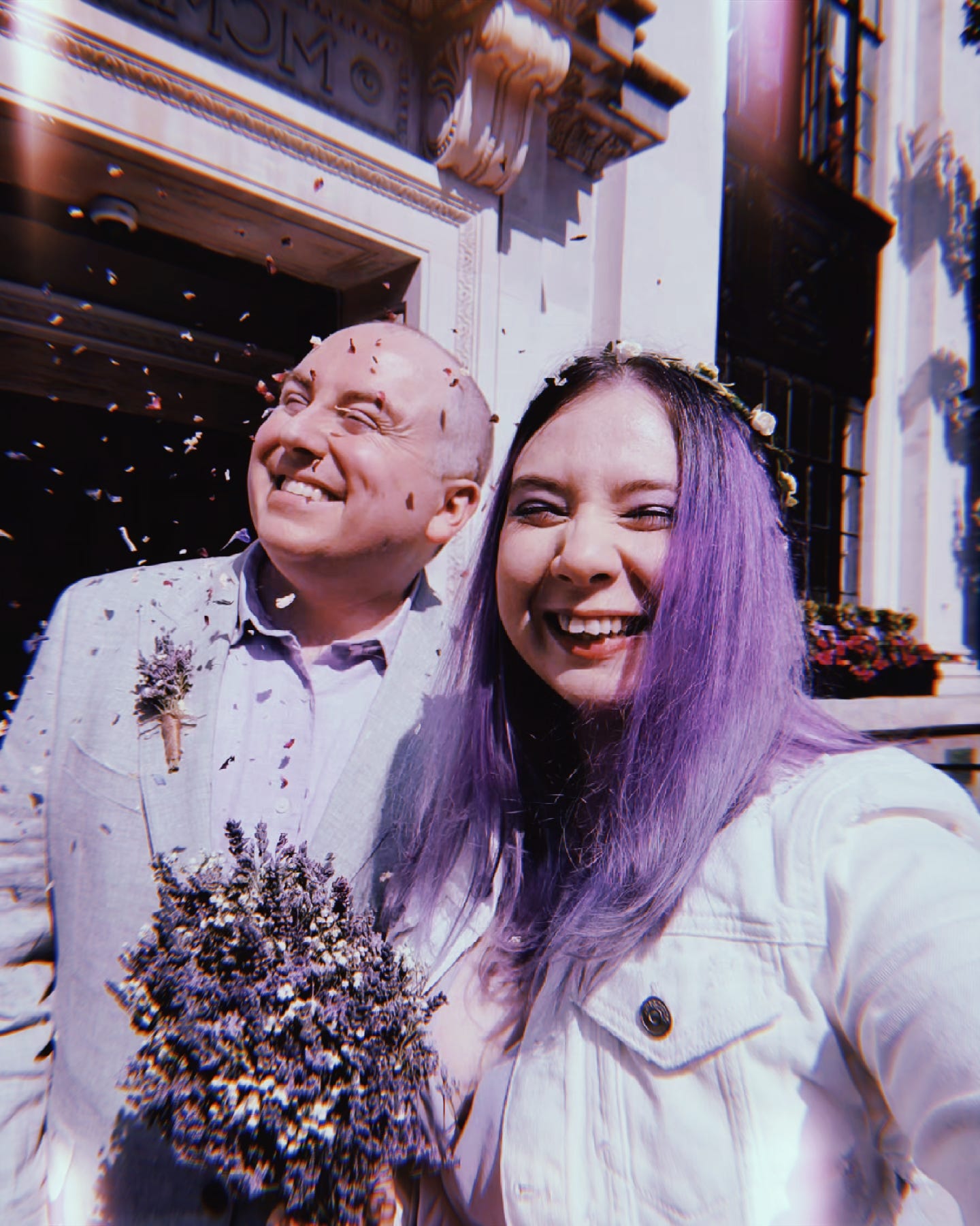 photo of a smiling couple getting flower petals thrown at them. kat's partner wears a gray linen suit with a lilac linen shirt and a dried lavender flower boutonniere. kat has long purple hair, and wears a fabric flower crown of small white roses wrapped around a green wreath, a white jean jacket, lilac dress, and holds a dried lavender flower bouquet.