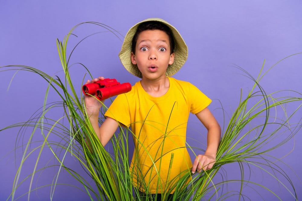 Young boy dressed in an explorer's hat, peeking out from behind a plant with red binoculars in his hand