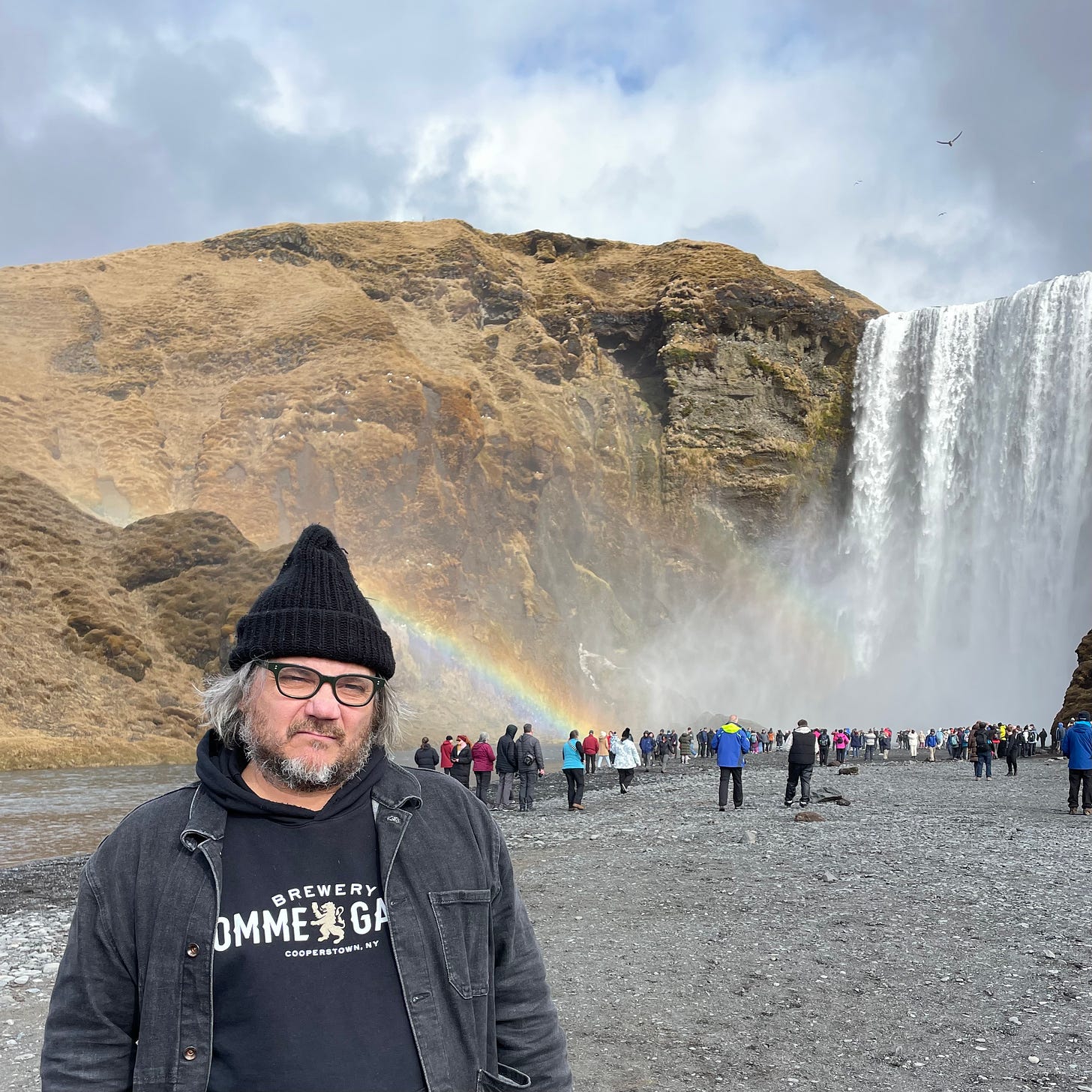 Jeff poses with a deadpan facial expression in front of a waterfall and a rainbow.