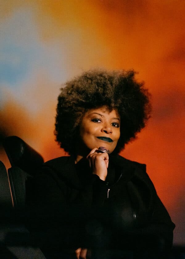 Ytasha Womack, wearing a black jacket and black lipstick, sits and smiles against an orange backdrop.