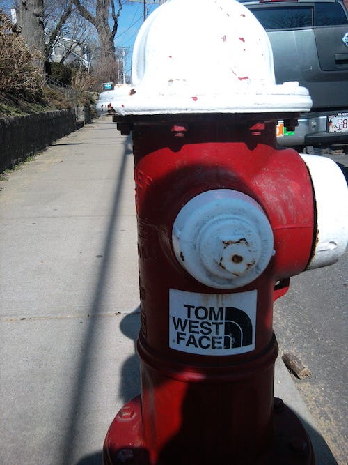 Photo of a fire hydrant with a sticker on it, a parody of the North Face logo, with the words 'Tom West Face'.