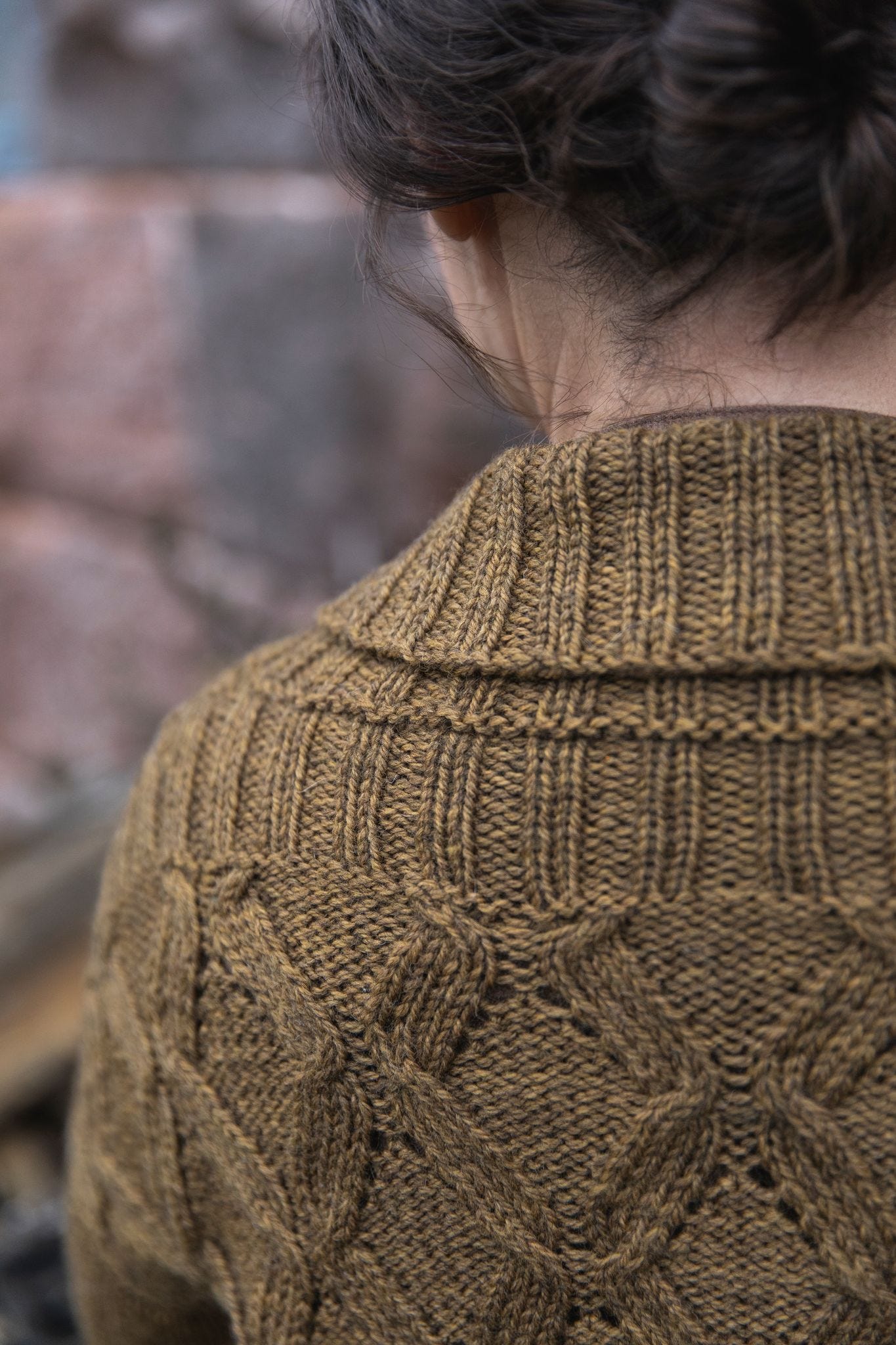 A partial view of a woman's back. Her dark hair is pulled up and she looks down to the left. Her cardigan features a cabled criss-cross motif and a very wide ribbed collar that is folded over. The sweater is a dark bronze color.