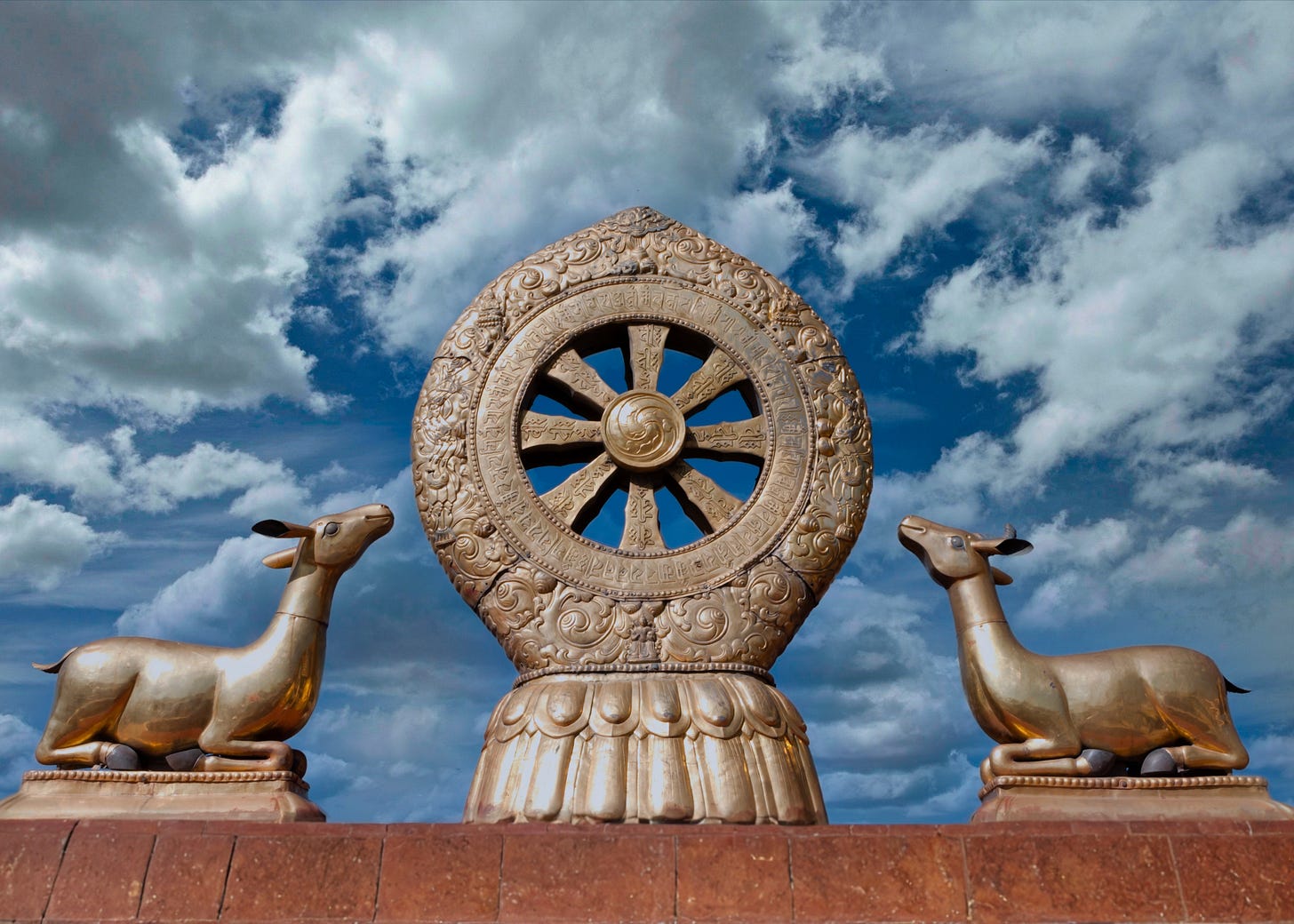 Sculpture of wheel with eight spokes with a dear on each side. Cloudy sky in background.