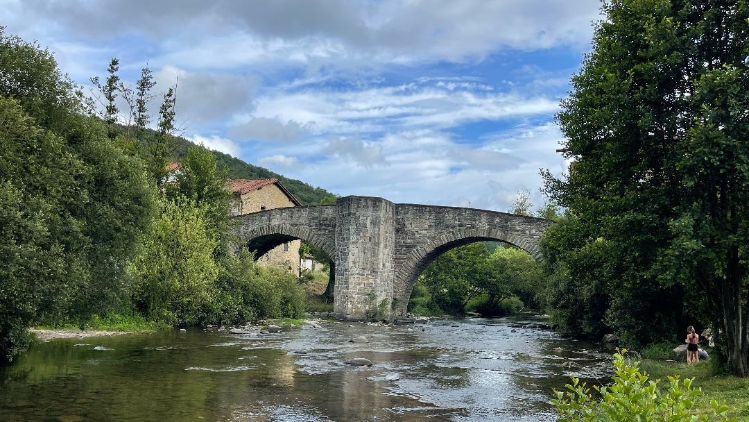 An ancient stone bridge with high arches over a tranquil river that you see in the foreground. It’s a sunny day, a blue sky with some smaller clouds. Left and right of the river are green trees.