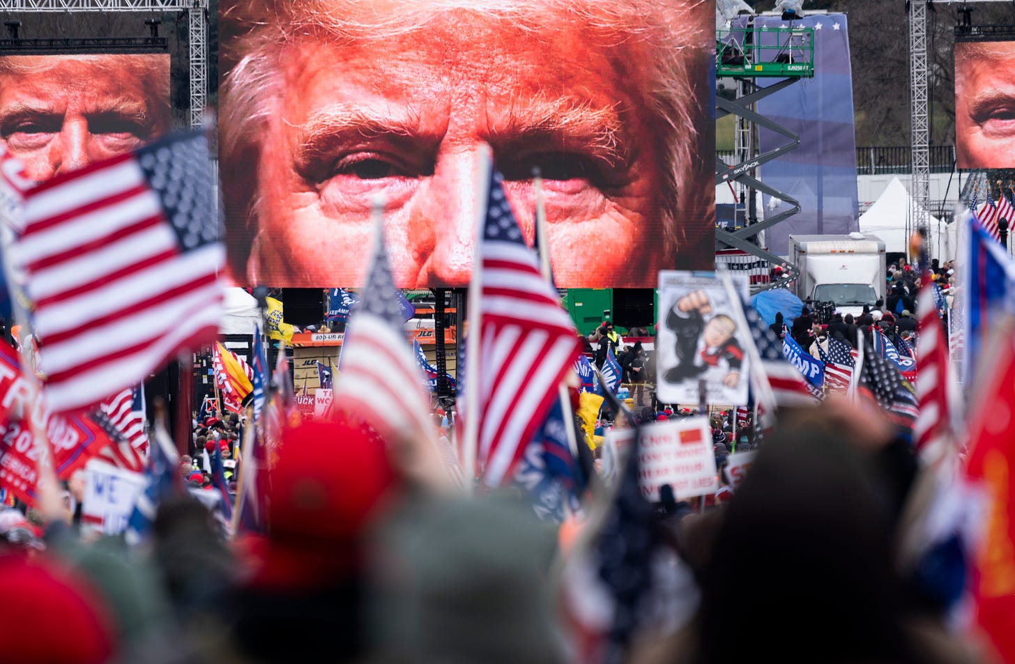 A photo of a large rally of people waving American flags and Trump signs, above the rally a large screen shows a closeup of the eyes of former president Donald Trump