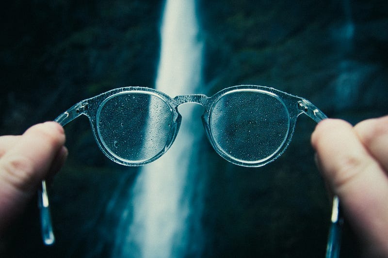 Person holding up glasses/spectacles with a blurred waterfall in the background.