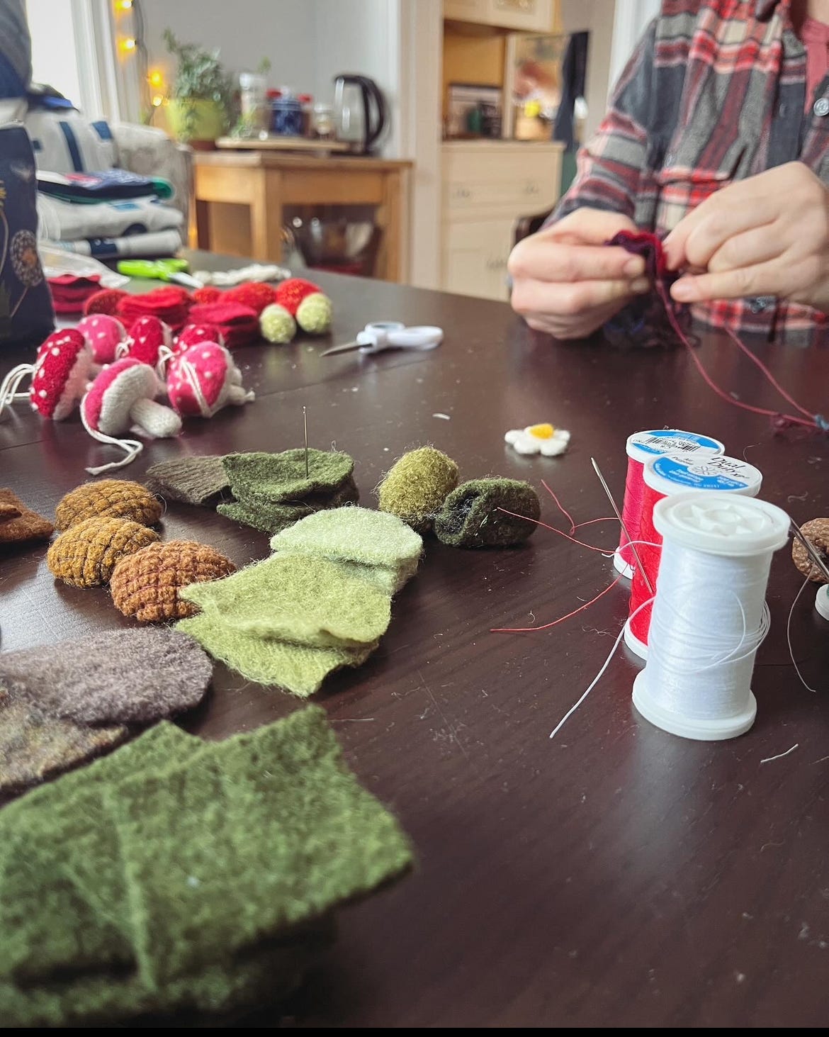 A dining room table covered with crafting supplies, including bits of felt, spools of thread, and some already finished crafts, including tiny felt mushroom ornaments.
