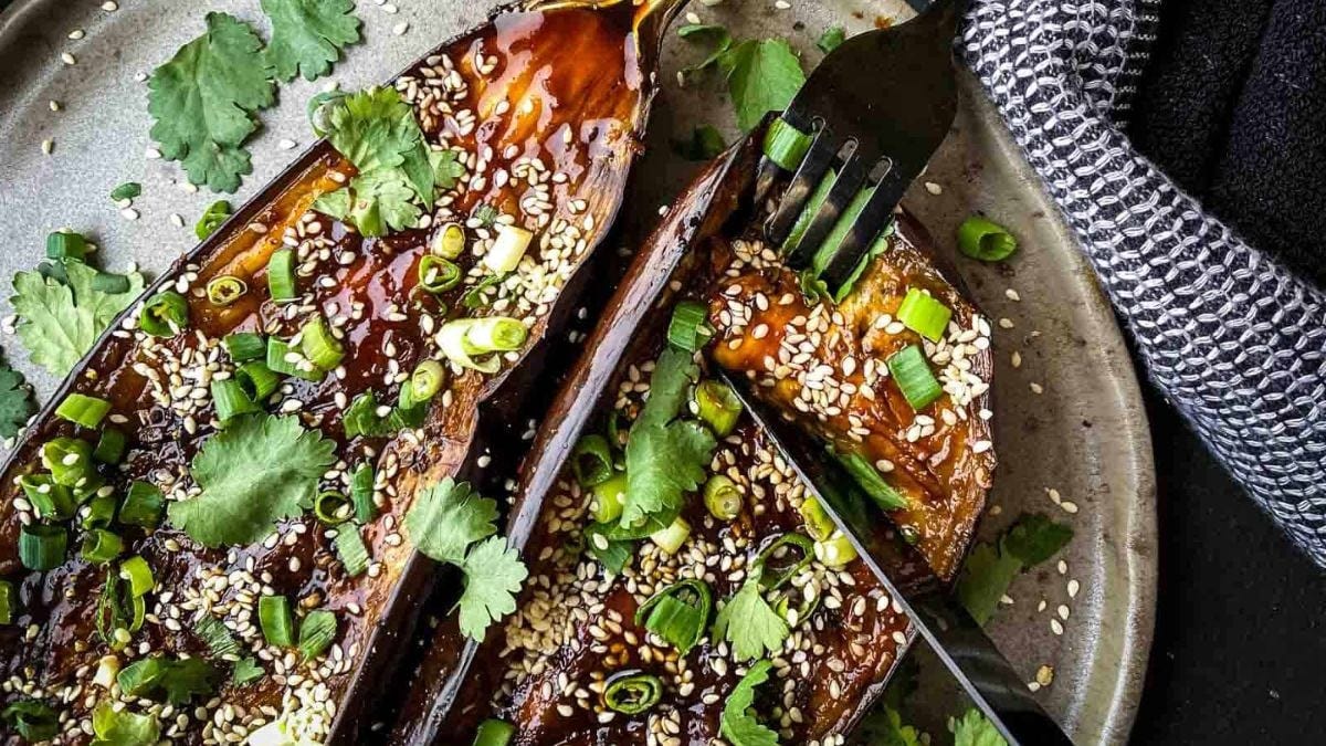 Grilled eggplants topped with sesame seeds and scallions, garnished with cilantro, served on a rustic plate with a fork on the side.
