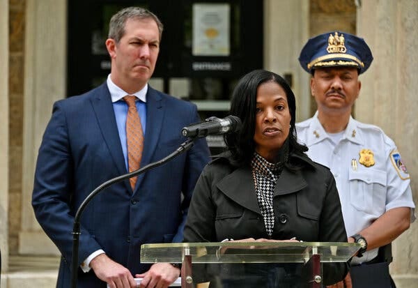 Myriam Rogers is speaking behind a lectern with a microphone pointing toward her. A police officer and another man in a suit are standing behind her.