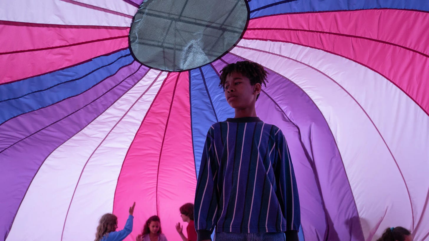 A prepubescent boy (Owen) stands under a parachute dome that is colored in stripes of white, pink, blue, and purple. there are children playing with each other under the dome in the background, but he is standing on his own looking confused.