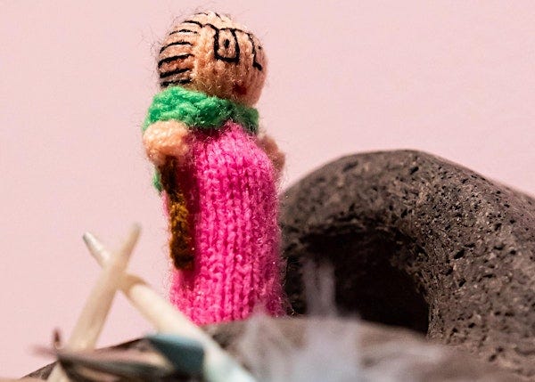 close up of a tiny knitted doll with square glasses and pink outfit