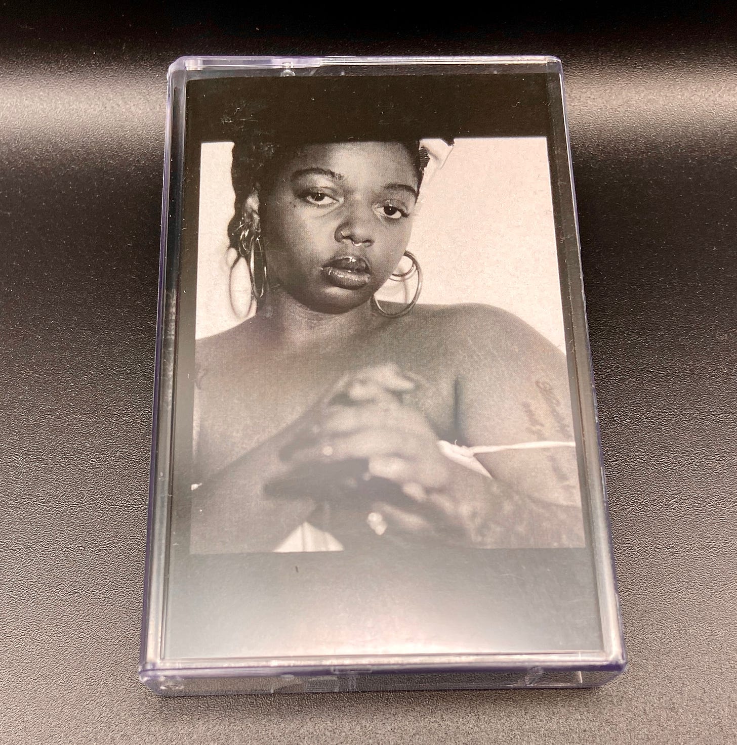 a cassette for keiyaA’s album Forever, Ya Girl. a Black woman glares at the camera with her hands clasped in front of her.