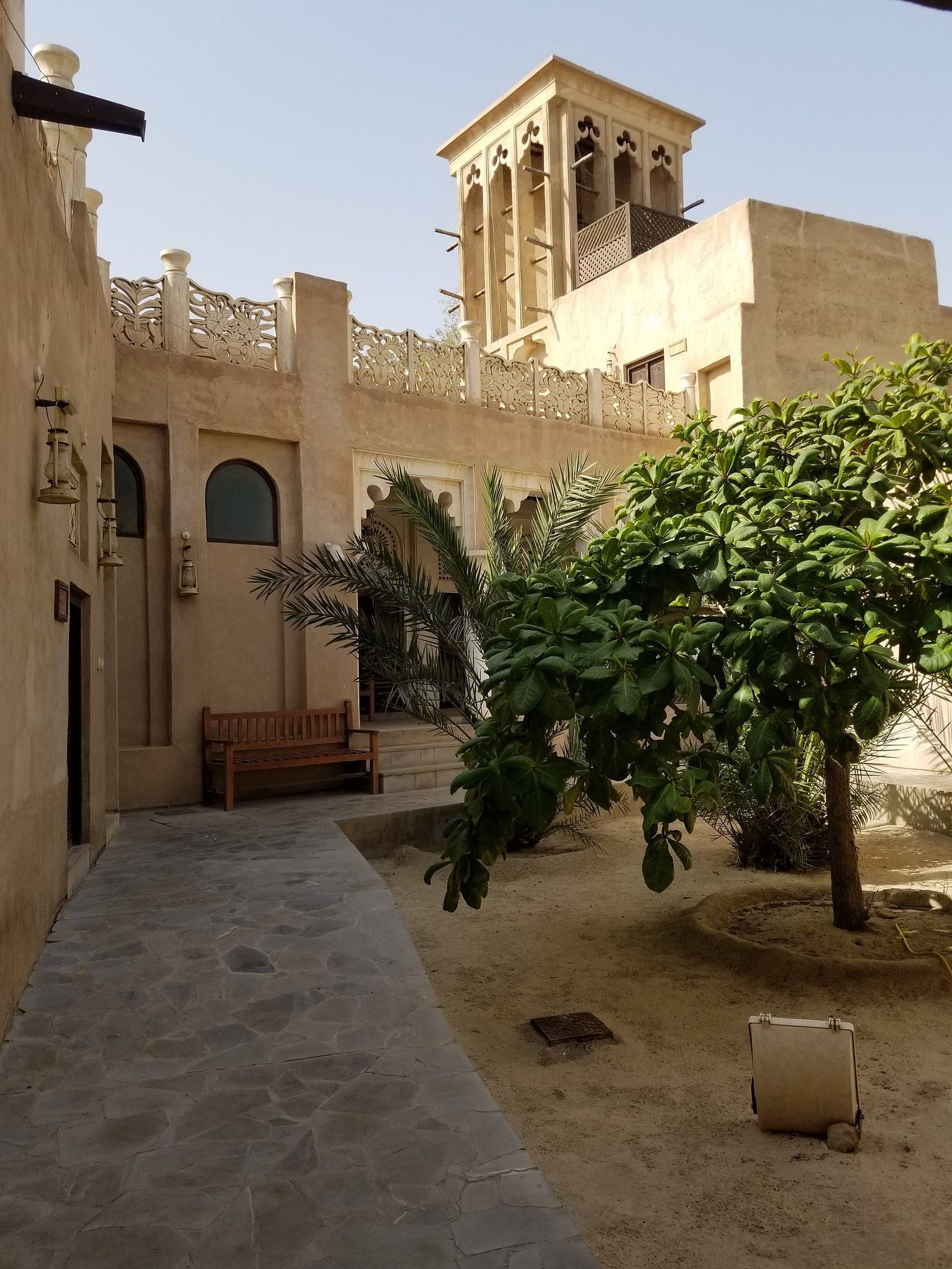 An afternoon photo of a historical Dubai-based courtyard with two trees and a wooden bench An afternoon photo of a historical Dubai-based courtyard with two trees and a wooden bench