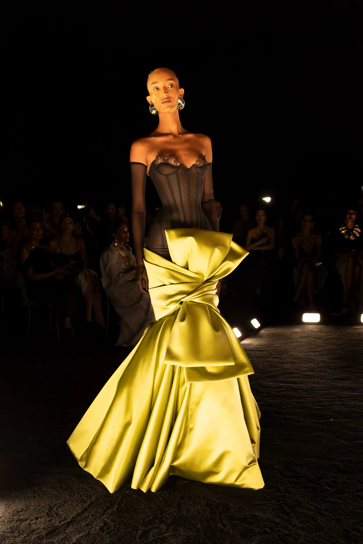 A dark-skinned model with slicked back hair walks a runway lit only by footlights wearing a gown with a sheer black corset top and a chartreuse satin skirt tied into a bow.