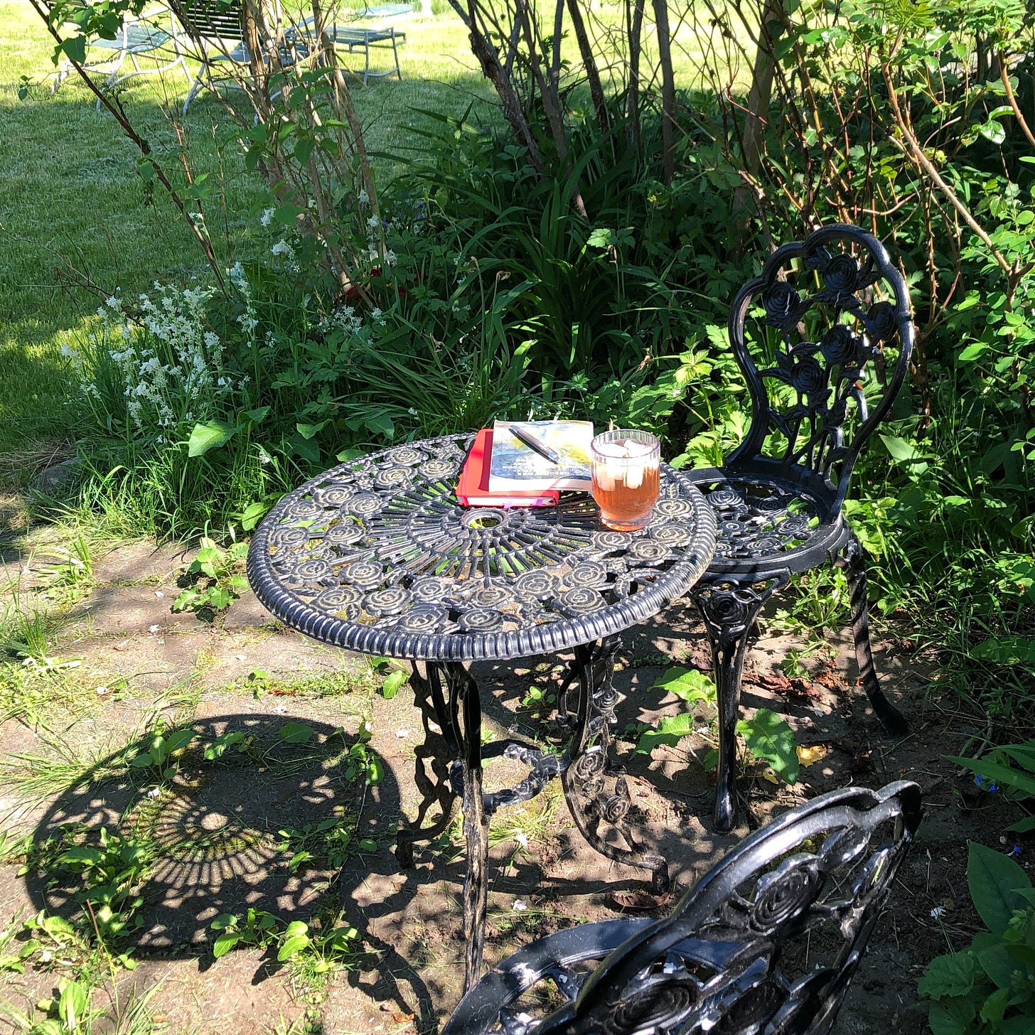 Black café-style metal chairs and table, holding book, notebook, pen, and iced tea, in a clearing surrounded by greenery.