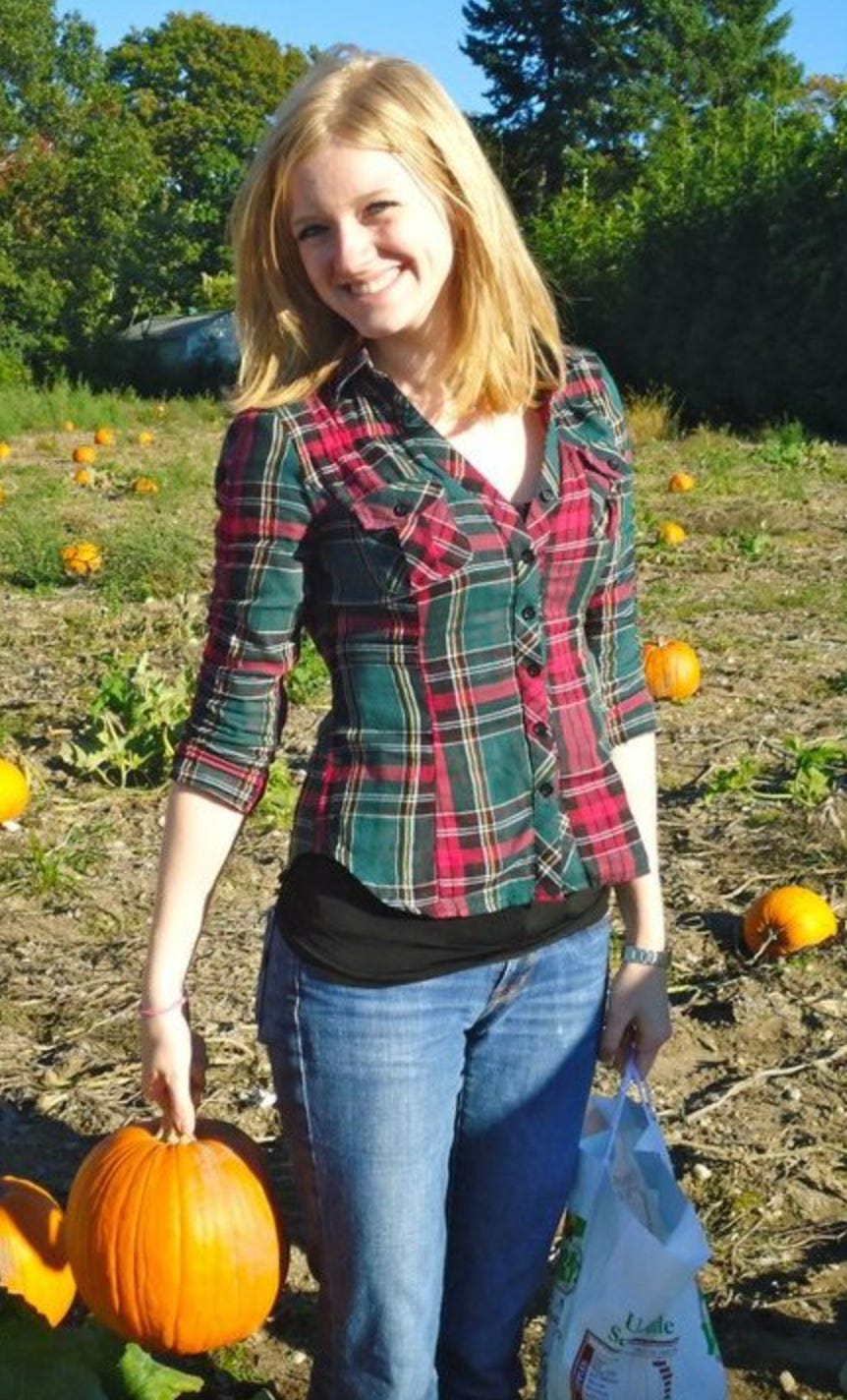 Carly standing in a pumpkin patch, wearing a red and green plaid shirt and holding a pumpkin.