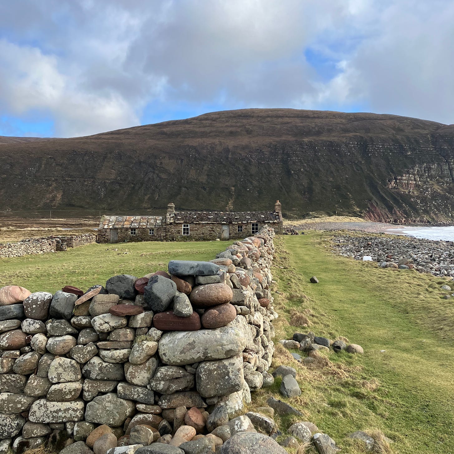 A stone house and low wall beside massive cliffs of Hoy on a sunny day with blue sky and clouds above.