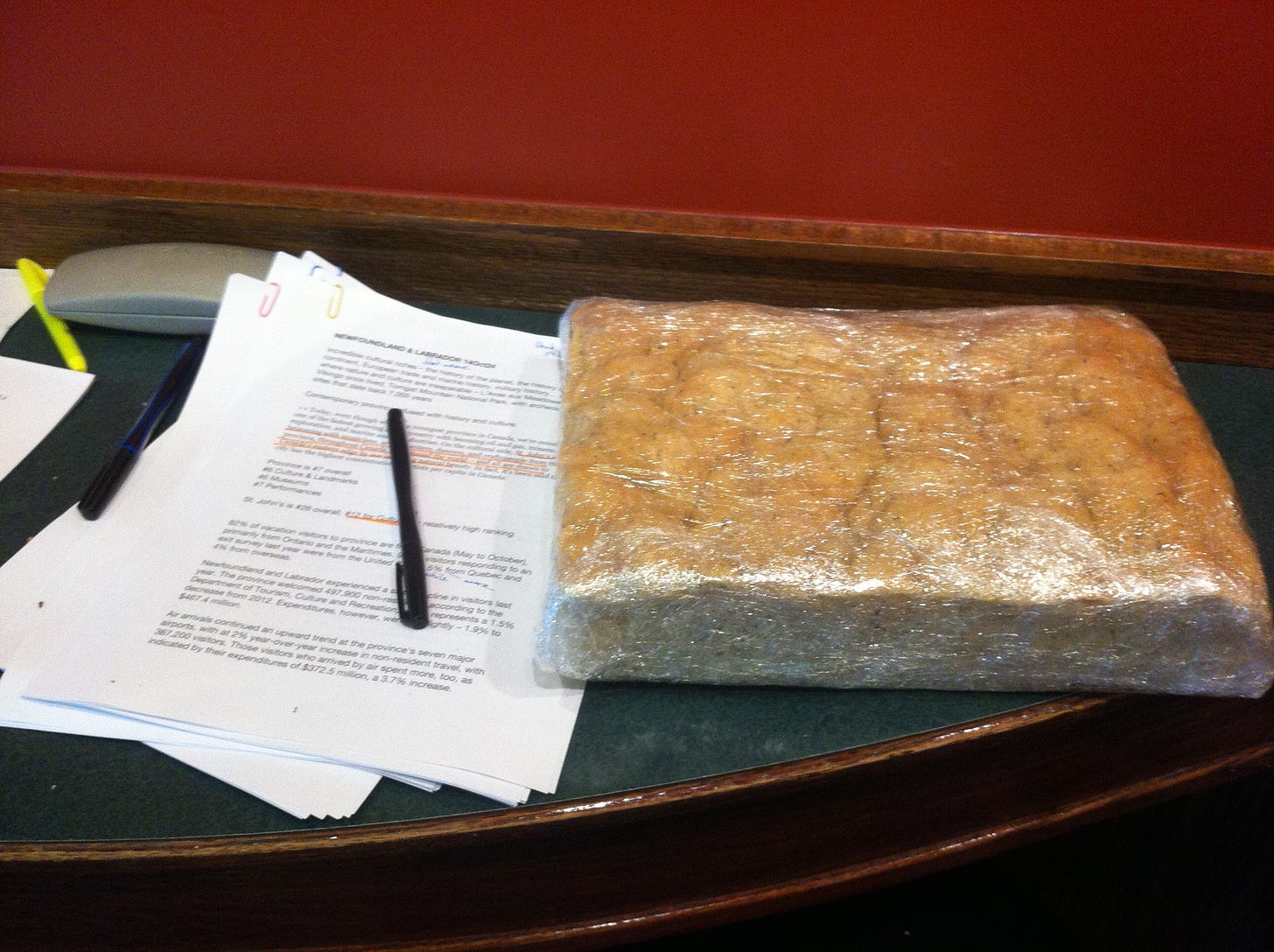 On the left, a stack of printouts with handwritten notes in red and a pen on top. On the right, a square of focaccia bread in plastic wrap.