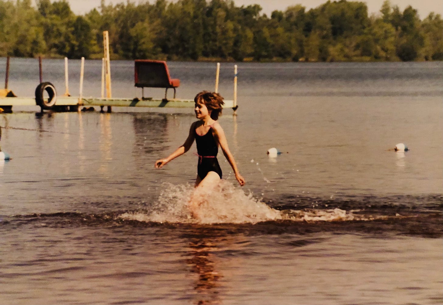 The author in a black swimsuit with a purple belt, running through the water inside the swim buoys with a dock, the lake and trees in the background.