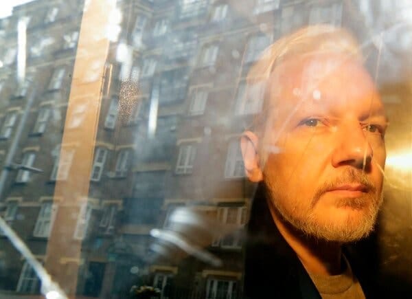 Julian Assange as seen through a window reflecting a building.
